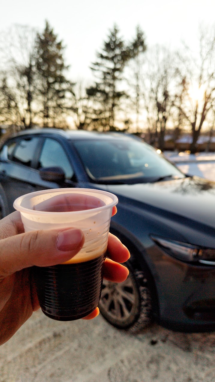 A hand holding a cup of espresso in front of a dark SUV on a snowy day at sunset.